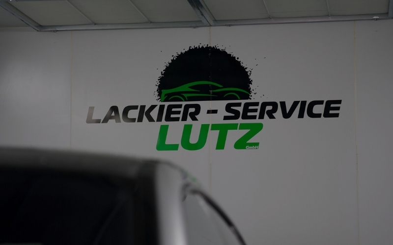 Eine Wand mit dem Schriftzug 'LACKIER - SERVICE LUTZ' in schwarzer und grüner Schrift, hinter einem unscharfen Fahrzeug. Der Schriftzug ist kombiniert mit einem grafischen Element eines Autos in der oberen Mitte der Wand.