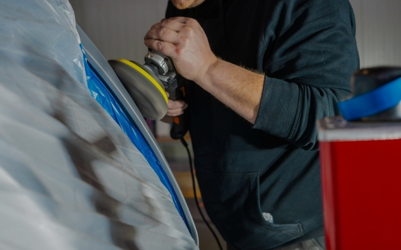 Ein Handwerker, der einen Polierer in der Hand hält und die Oberfläche eines Fahrzeugs bearbeitet, während das Fahrzeug mit einer blauen Abdeckfolie geschützt ist.