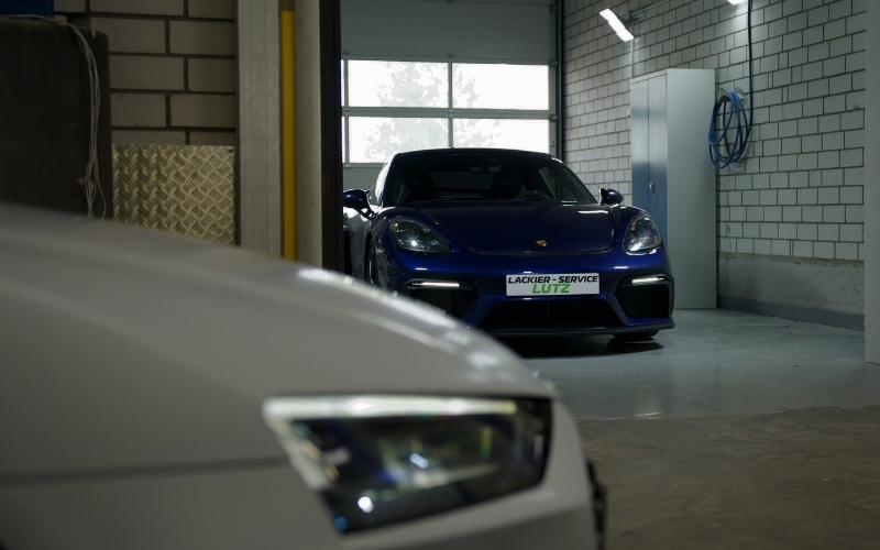 Eine blaue Porsche-Sportwagen steht in einer Werkstatt, während ein graues Fahrzeug im Vordergrund teilweise sichtbar ist und der Werkstattaufschrift "LACKIER-SERVICE LUTZ" im Hintergrund zu sehen ist.